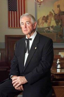 An older man in a black suit and string tie seated in a courtroom beside an American flag and a western mural